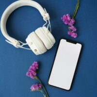 Stylish flat lay of white headphones and smartphone on blue surface with purple flowers.